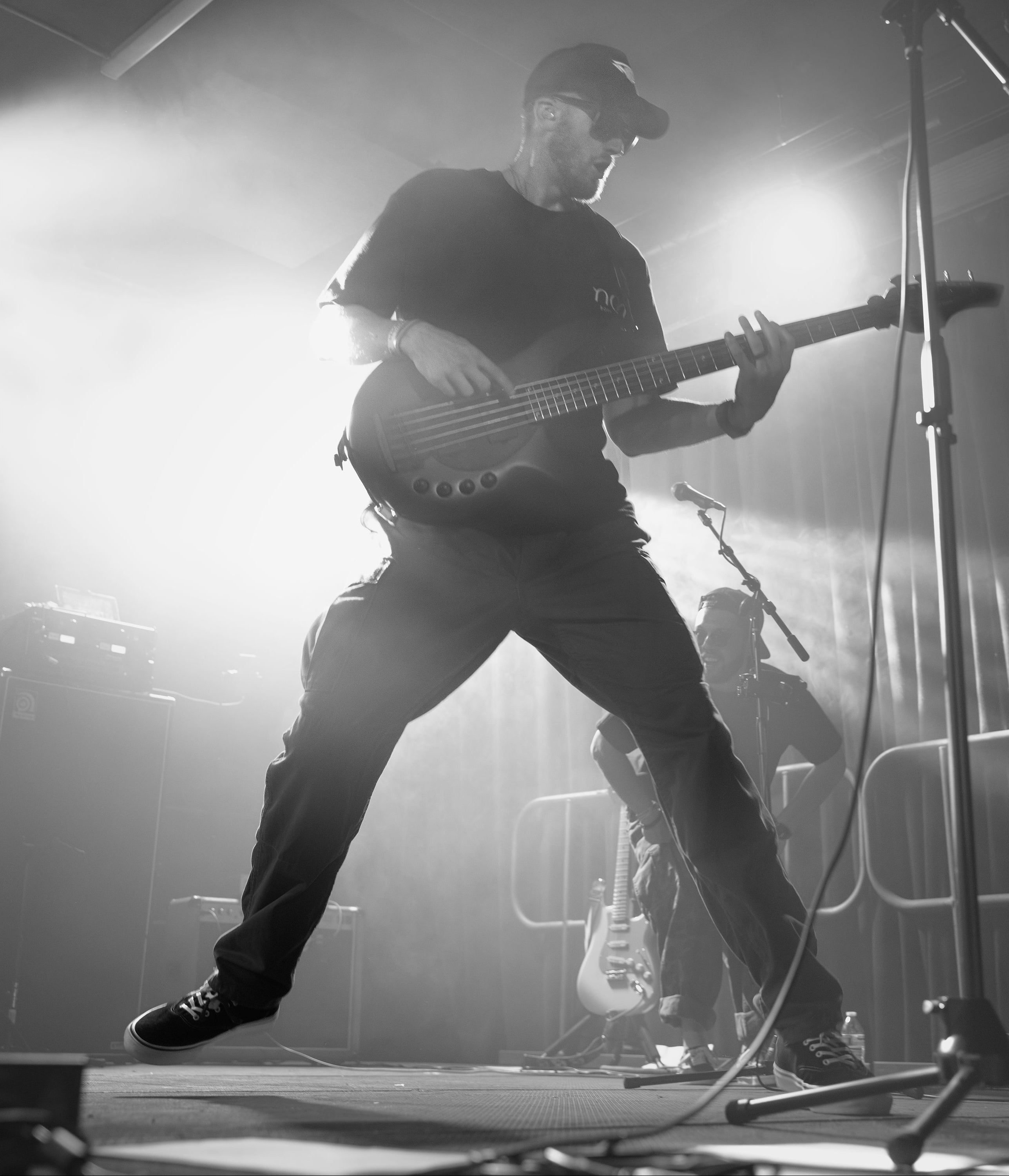 Person playing bass guitar on stage with a microphone stand and another person in the background.